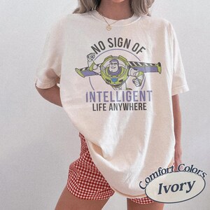 May include: Ivory-colored t-shirt featuring a graphic of Buzz Lightyear with the text "NO SIGN OF INTELLIGENT LIFE ANYWHERE." The shirt is a Comfort Colors brand. The model is wearing red and white checkered shorts.