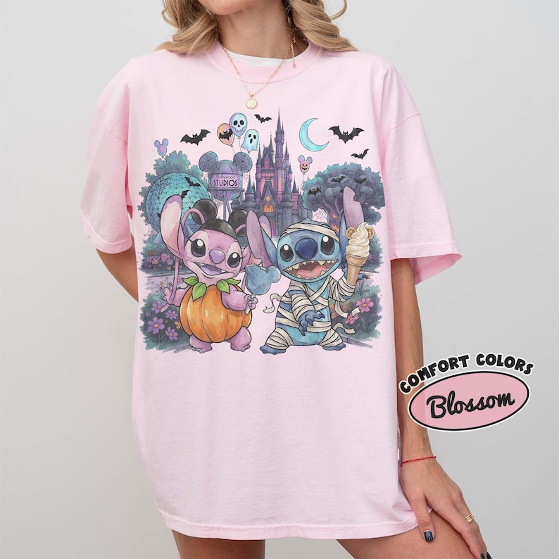 May include: A light pink t-shirt featuring a graphic print of two blue and pink alien characters in Halloween costumes, one dressed as a pumpkin and the other as a mummy, set against a backdrop of a castle, trees, bats, and a crescent moon.
