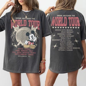 May include: Dark gray t-shirt with a graphic of Mickey Mouse in front of a geometric sphere, with the text "DRINK AROUNDE THE WORLD TOUR". The back of the shirt lists cities. A fun, vintage-style tee.