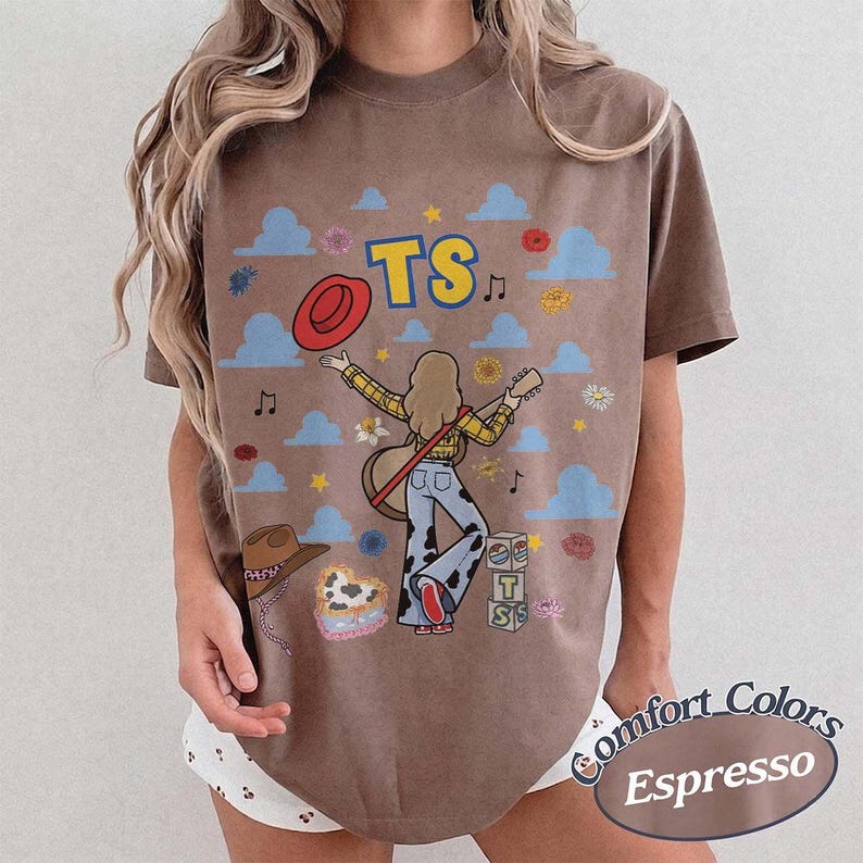 May include: A brown short-sleeve t-shirt featuring a graphic of a person with long hair playing a guitar, wearing a yellow plaid shirt and cow-print pants. The design includes blue clouds, flowers, a red hat, and blocks with the letters TS.