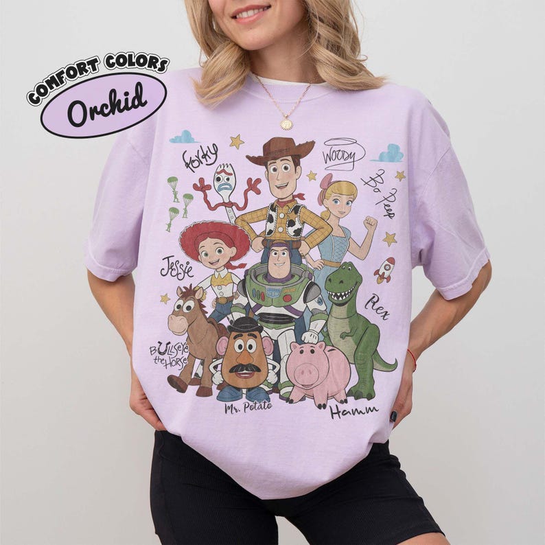 May include: An orchid-colored t-shirt featuring a graphic print of Toy Story characters including Woody, Buzz Lightyear, Jessie, Rex, Hamm, Mr. Potato Head, Bullseye, Bo Peep, and Forky, with their names written in a cursive script font.
