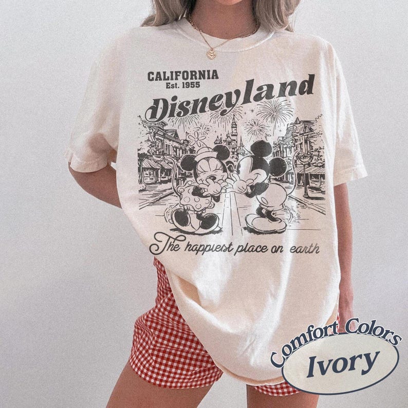 Puede incluir: Camiseta de color marfil con un gr&aacute;fico de Disneyland con Mickey y Minnie Mouse. El gr&aacute;fico incluye el texto "CALIFORNIA Est. 1955" y "The happiest place on earth". La camiseta se combina con pantalones cortos a cuadros rojos y blancos.