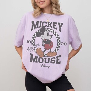 May include: A light purple t-shirt featuring a distressed graphic of Mickey Mouse. The design includes the words "Mickey Mouse" and "Disney" along with the years "1928" and "19". The shirt has short sleeves and a relaxed fit.