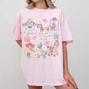 K&ouml;nnte beinhalten: Ein hellrosa T-Shirt mit einer bunten Grafik von Zeichentrickfiguren, darunter ein Weltraumranger, ein Cowboy und ein gr&uuml;ner Dinosaurier. Der Text "YOU'VE GOT A FRIEND IN ME" ist ebenfalls sichtbar.