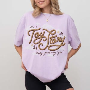 May include: A light purple short-sleeve t-shirt featuring a graphic print of the text "it's a Toy Story" in a brown rope-like font, accompanied by small clouds, a flower, and the additional text "Baby, just say "yes"" in a dark brown script.