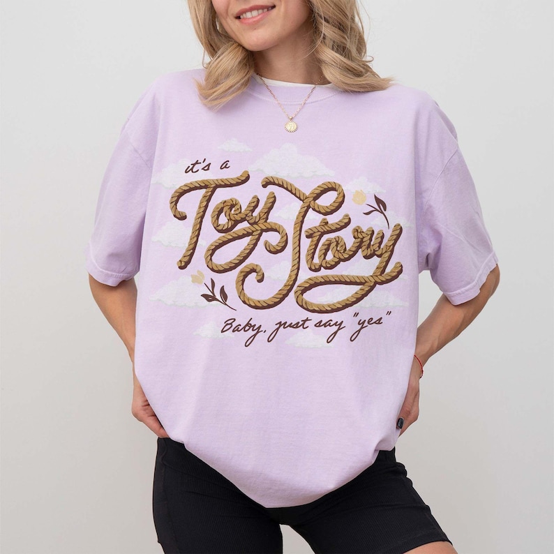 May include: A light purple short-sleeve t-shirt featuring a graphic print of the text "it's a Toy Story" in a brown rope-like font, accompanied by small clouds, a flower, and the additional text "Baby, just say "yes"" in a dark brown script.