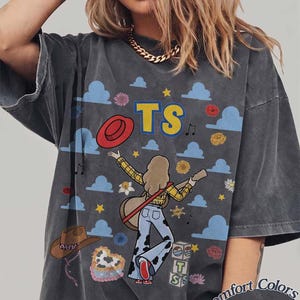 May include: A charcoal gray t-shirt featuring a graphic print of a person with long hair playing an acoustic guitar. The design includes blue clouds, yellow stars, a red hat, a brown cowboy hat, and the letters TS. Text at the bottom reads Comfort Colors Pepper.