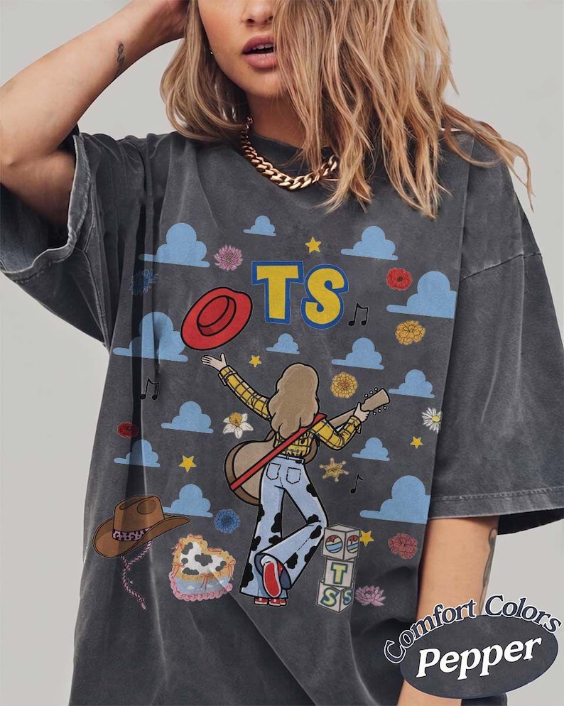 May include: A charcoal gray t-shirt featuring a graphic print of a person with long hair playing an acoustic guitar. The design includes blue clouds, yellow stars, a red hat, a brown cowboy hat, and the letters TS. Text at the bottom reads Comfort Colors Pepper.