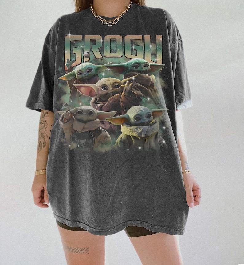 Puede incluir: Camiseta gris oscuro con la palabra "GROGU" en una fuente retro colorida. La camiseta muestra m&uacute;ltiples im&aacute;genes de Grogu, tambi&eacute;n conocido como Baby Yoda, de la serie Star Wars. El dise&ntilde;o est&aacute; impreso en una camiseta suave y de gran tama&ntilde;o.