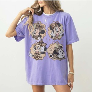 May include: A lilac-colored t-shirt featuring four illustrations of Minnie Mouse in leopard print outfits. Each illustration is framed in a tan-colored oval with gold accents. The shirt has short sleeves and a relaxed fit.