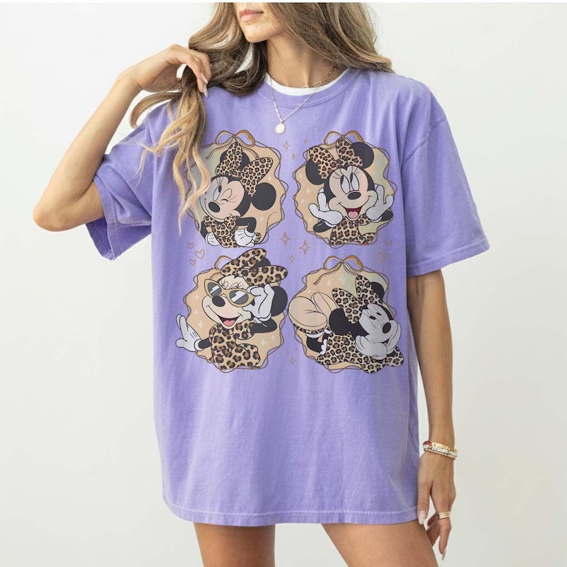 May include: A lilac-colored t-shirt featuring four illustrations of Minnie Mouse in leopard print outfits. Each illustration is framed in a tan-colored oval with gold accents. The shirt has short sleeves and a relaxed fit.