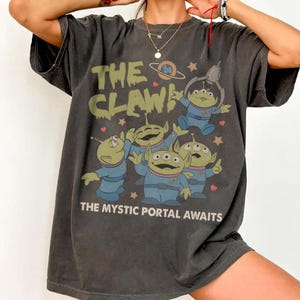 May include: Dark gray t-shirt featuring a graphic of green aliens in blue suits with the text "THE CLAW" and "THE MYSTIC PORTAL AWAITS". The aliens are in various poses, with stars and hearts around them.