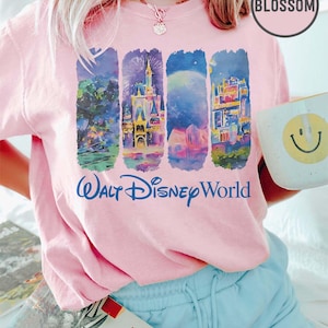 May include: A light pink t-shirt featuring four vertical panels with colorful illustrations of Walt Disney World scenes. The words "Walt Disney World" are printed in blue script below the panels. A white mug with a yellow smiley face is held in the hand.