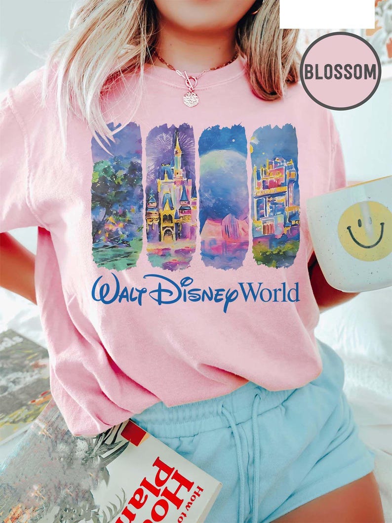 May include: A light pink t-shirt featuring four vertical panels with colorful illustrations of Walt Disney World scenes. The words "Walt Disney World" are printed in blue script below the panels. A white mug with a yellow smiley face is held in the hand.