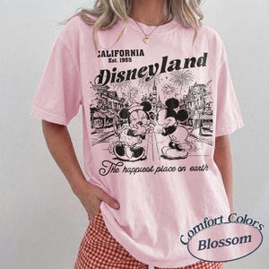 Puede incluir: Camiseta rosa claro con un gr&aacute;fico de Disneyland en blanco y negro con Mickey y Minnie Mouse. La camiseta incluye el texto "California Est. 1955" y "The happiest place on earth". La camiseta es un Comfort Colors Blossom.