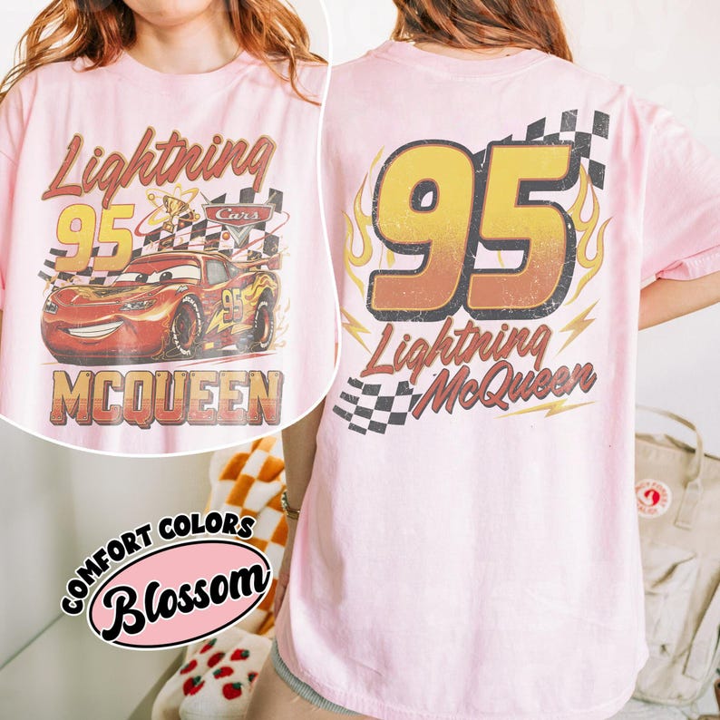 Może przedstawiać: R&oacute;żowa koszulka z grafiką Zygzaka McQueena z filmu Auta. Prz&oacute;d koszulki zdobi napis "Lightning 95 McQueen" z szachownicą i postacią z kresk&oacute;wki. Tył koszulki przedstawia napis "95 Lightning McQueen".