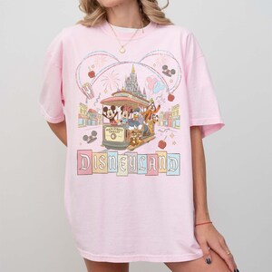 May include: Pink t-shirt featuring a colorful Disneyland graphic. The design includes a trolley with Mickey Mouse, Donald Duck, and Goofy, along with the Disneyland logo. The graphic is surrounded by fireworks, hearts, and other decorative elements.
