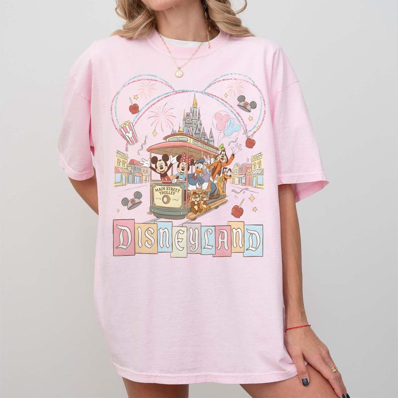 May include: Pink t-shirt featuring a colorful Disneyland graphic. The design includes a trolley with Mickey Mouse, Donald Duck, and Goofy, along with the Disneyland logo. The graphic is surrounded by fireworks, hearts, and other decorative elements.