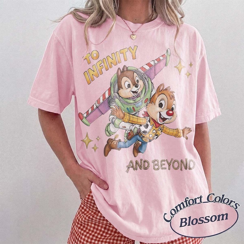 Puede incluir: Camiseta rosa claro con un dise&ntilde;o de Chip y Chop con un traje espacial inspirado en Buzz Lightyear y un disfraz de Woody, con el texto "TO INFINITY AND BEYOND." La camiseta es de Comfort Colors.