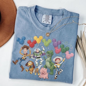May include: A blue t-shirt featuring a graphic of Toy Story characters including Woody, Buzz Lightyear, Rex, Slinky Dog, Hamm, and Jessie. The characters are surrounded by colorful Mickey Mouse-shaped balloons. The shirt has a Comfort Colors label.