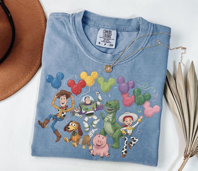 May include: A blue t-shirt featuring a graphic of Toy Story characters including Woody, Buzz Lightyear, Rex, Slinky Dog, Hamm, and Jessie. The characters are surrounded by colorful Mickey Mouse-shaped balloons. The shirt has a Comfort Colors label.