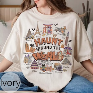 Puede incluir: Camiseta de color marfil con un diseño temático de Halloween. El diseño presenta el texto "Haunt Around The World" con ilustraciones de monumentos de varios países, como la Torre Eiffel, el Big Ben y la Estatua de la Libertad.
