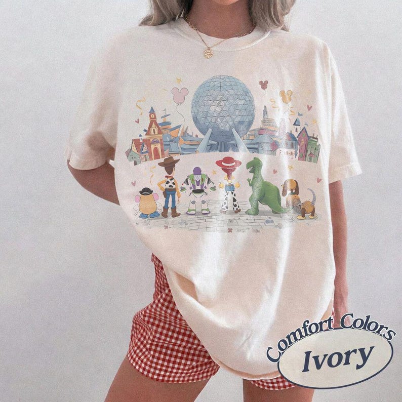 Puede incluir: Camiseta de color marfil con un gr&aacute;fico colorido de personajes de Disney y un parque tem&aacute;tico. El dise&ntilde;o incluye una gran esfera azul, edificios y personajes de dibujos animados. La camiseta tiene mangas cortas y un corte holgado.