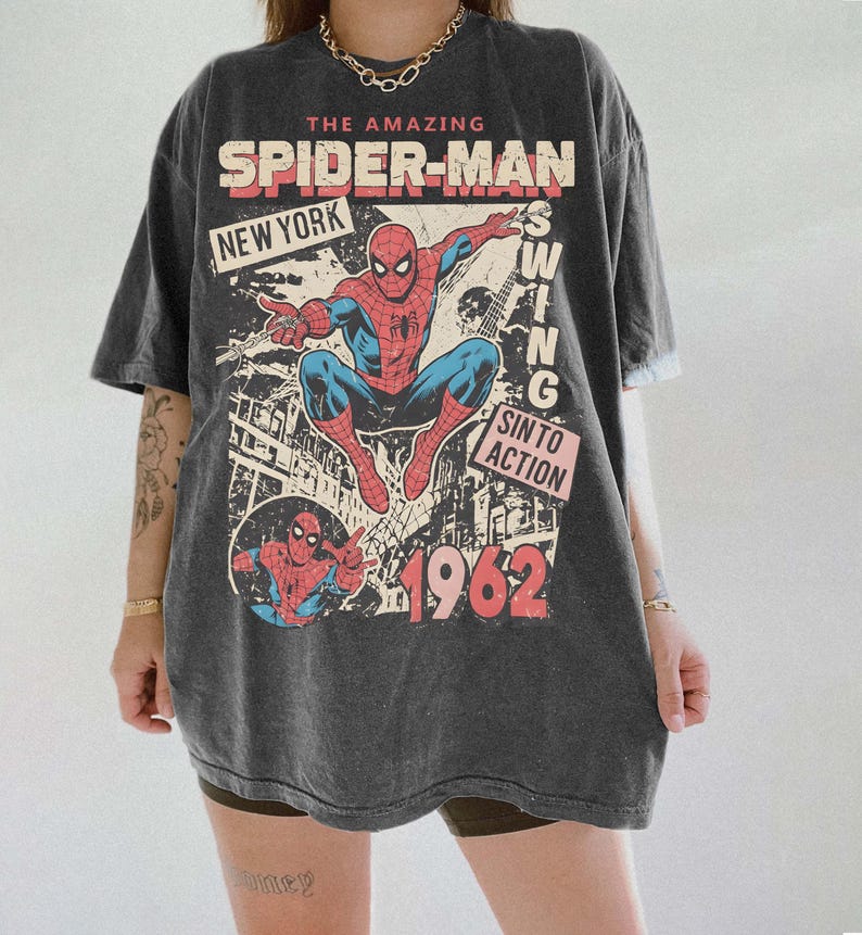 K&ouml;nnte beinhalten: Dunkelgraues T-Shirt mit einem Vintage-Comic-Design von Spider-Man. Die Grafik enth&auml;lt den Text "The Amazing Spider-Man", "New York" und das Jahr "1962". Der Superheld ist in seinem klassischen rot-blauen Anzug dargestellt.