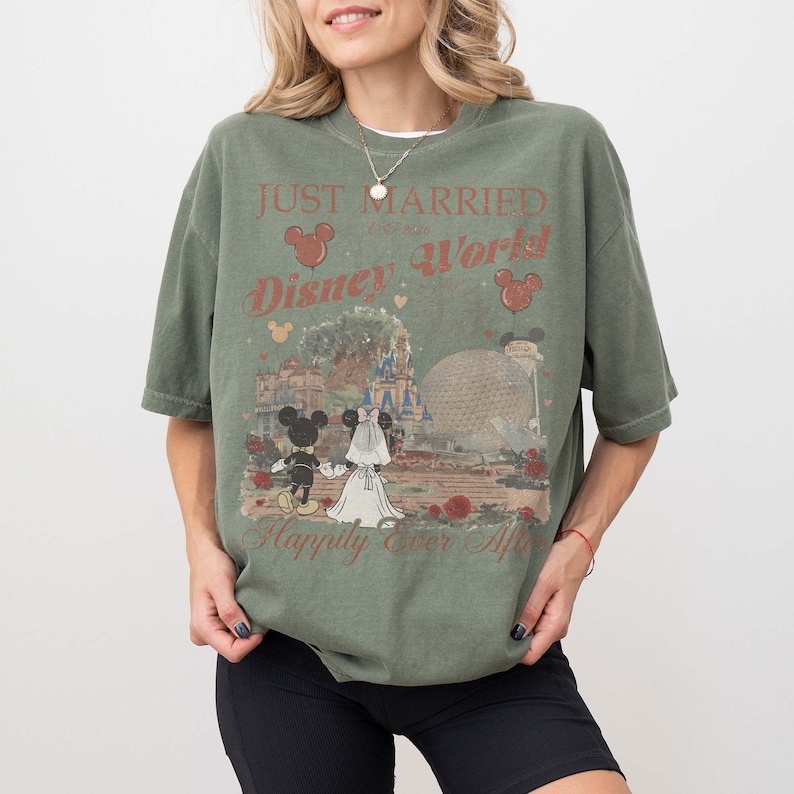 Pu&ograve; includere: T-shirt verde oliva con grafica Disney World. Il design presenta Topolino e Minnie in abiti da sposi, con la scritta "Just Married" e "Happily Ever After". La maglietta ha uno stile vintage.