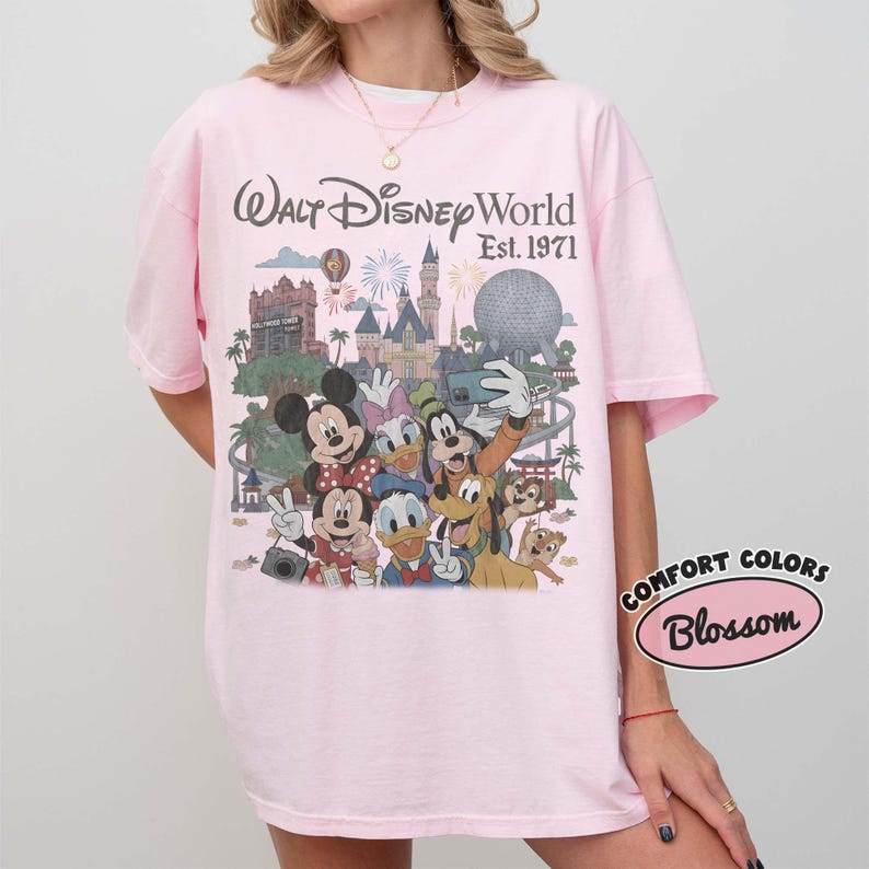 May include: A light pink t-shirt featuring a graphic print of Walt Disney World Est. 1971 with various cartoon characters including Mickey Mouse, Minnie Mouse, Donald Duck, Daisy Duck, Goofy, Pluto, and Chip 'n Dale in front of park landmarks.