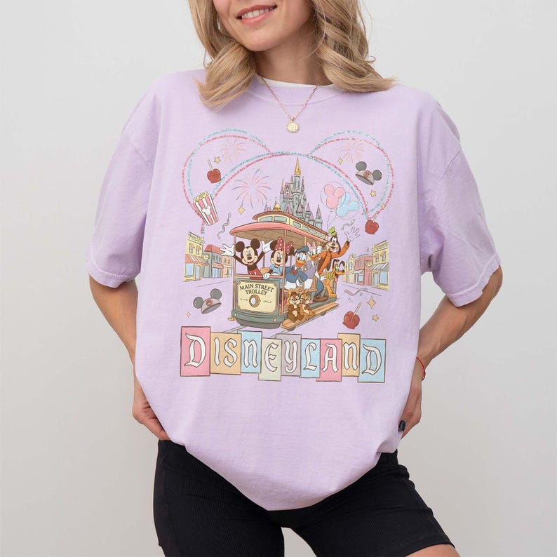 May include: A light purple t-shirt featuring a Disneyland design. The graphic includes a trolley with Mickey Mouse, Donald Duck, and Goofy, with the word "Disneyland" in colorful letters. The design is framed by a Mickey Mouse head shape.