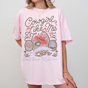 May include: A light pink short-sleeve t-shirt featuring a brown rope-style graphic with the text "Cowgirl Like Me" and "Takes One to Know One." The design includes a red cowboy hat, a horseshoe, roses, musical notes, and small stars.