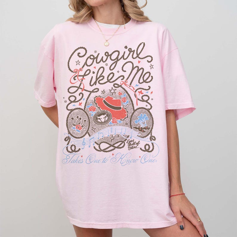 May include: A light pink short-sleeve t-shirt featuring a brown rope-style graphic with the text "Cowgirl Like Me" and "Takes One to Know One." The design includes a red cowboy hat, a horseshoe, roses, musical notes, and small stars.