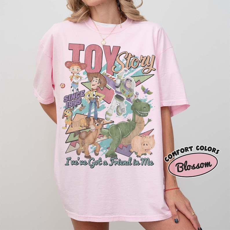May include: A light pink t-shirt featuring a graphic print of Toy Story characters including Woody, Buzz Lightyear, Jessie, Rex, Slinky Dog, Bullseye, and Hamm. The text reads TOY Story SINCE 1995 and I've Got a Friend in Me. A Comfort Colors Blossom label is visible.