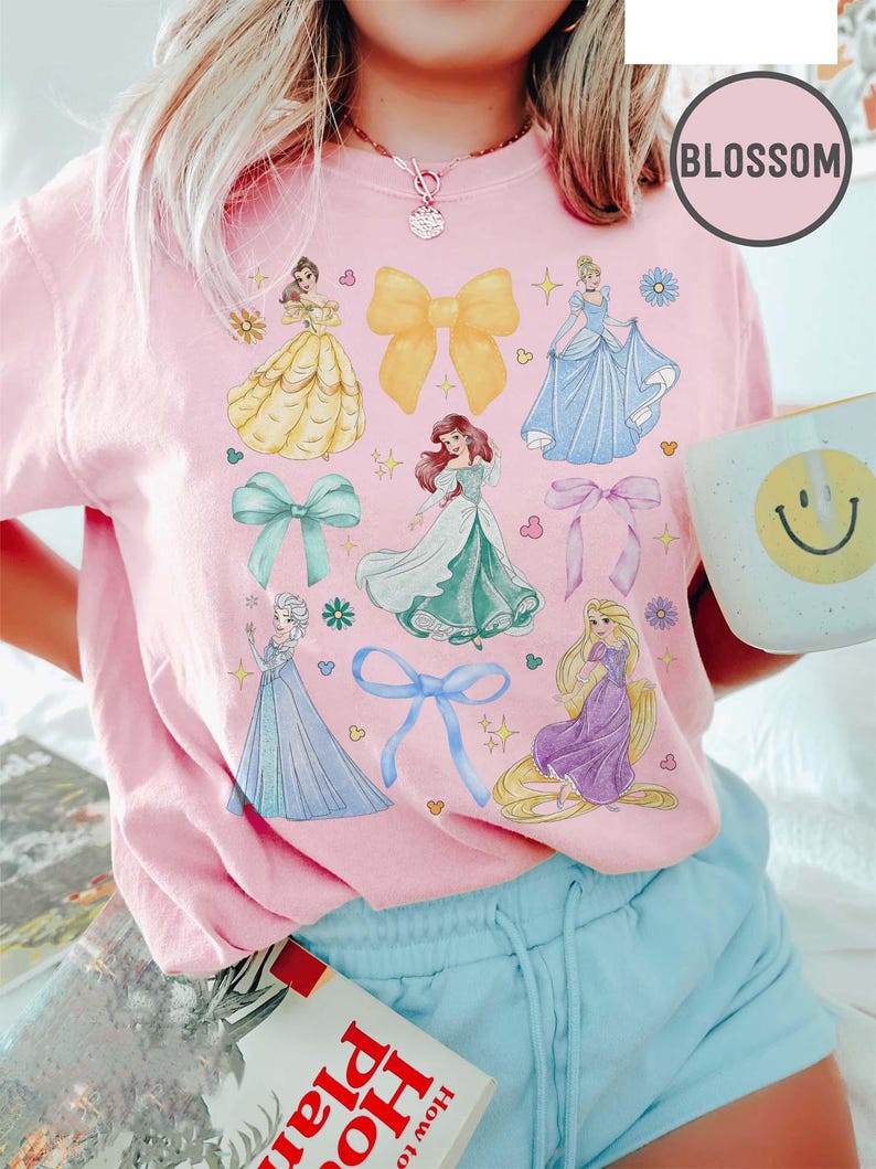 May include: Pink t-shirt featuring illustrations of princesses and bows. The shirt has a crew neck and short sleeves. The word "BLOSSOM" is printed on the shirt. The shirt is paired with light blue shorts.