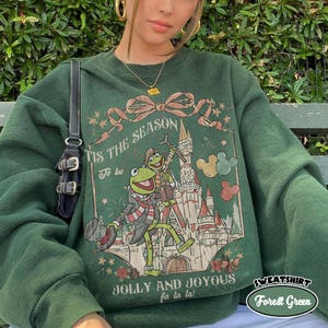 May include: A forest green crewneck sweatshirt featuring a graphic print of a frog character in a scarf standing before a castle. The text reads 'Tis the season' and 'Jolly and joyous fa la la'. A large ribbon bow sits above the castle illustration.