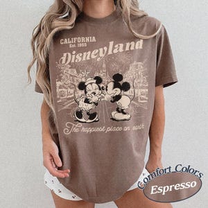 Puede incluir: Camiseta color espresso con un gr&aacute;fico de Disneyland. El dise&ntilde;o incluye el texto "California Est. 1955 Disneyland" sobre una ilustraci&oacute;n de Mickey y Minnie Mouse. Tambi&eacute;n est&aacute; presente la frase "The happiest place on earth".