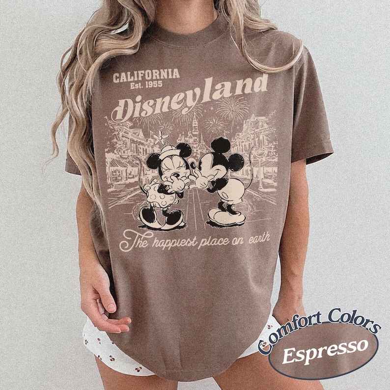 Puede incluir: Camiseta color espresso con un gr&aacute;fico de Disneyland. El dise&ntilde;o incluye el texto "California Est. 1955 Disneyland" sobre una ilustraci&oacute;n de Mickey y Minnie Mouse. Tambi&eacute;n est&aacute; presente la frase "The happiest place on earth".
