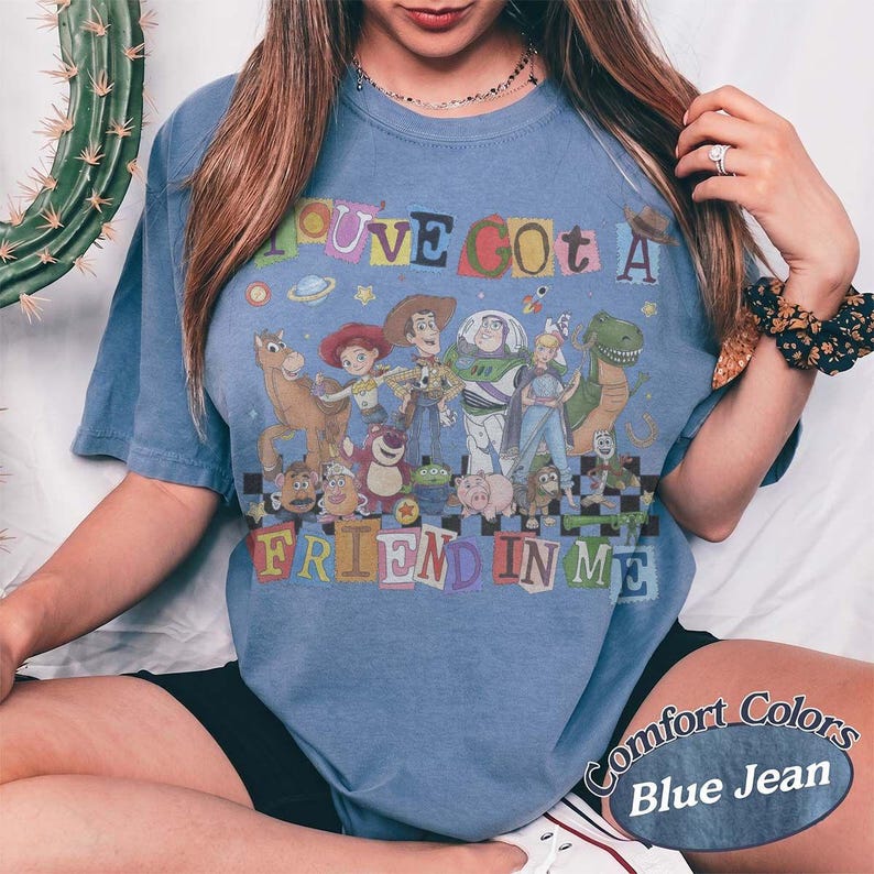 May include: A blue denim-colored t-shirt featuring a graphic print of various Toy Story characters. The text "YOU'VE GOT A FRIEND IN ME" is displayed in colorful, blocky letters above and below the characters, set against a checkered pattern.