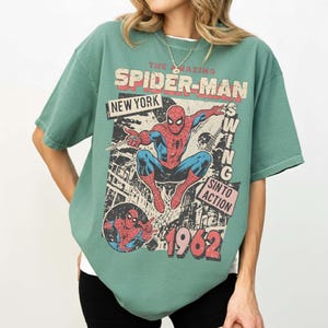 K&ouml;nnte beinhalten: Gr&uuml;nes T-Shirt mit einem Spider-Man-Motiv. Der Aufdruck zeigt "THE AMAZING SPIDER-MAN NEW YORK" und "SIN TO ACTION 1962". Das Shirt hat kurze &Auml;rmel und eine lockere Passform f&uuml;r einen l&auml;ssigen Look.