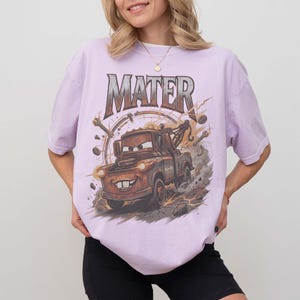 Puede incluir: Camiseta lila claro con una imagen de dibujos animados de Mate de la pel&iacute;cula Cars. El gr&aacute;fico incluye el nombre "MATER" en una fuente desgastada sobre el cami&oacute;n. El dise&ntilde;o tiene un aspecto vintage y desgastado.