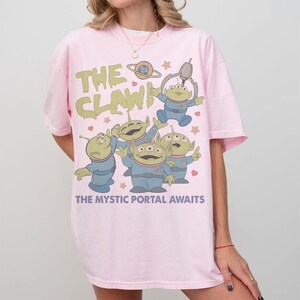 May include: Light pink t-shirt featuring a cartoon graphic of the aliens from the movie Toy Story. The graphic includes the text "THE CLAW!" and "THE MYSTIC PORTAL AWAITS". The aliens are in blue uniforms.