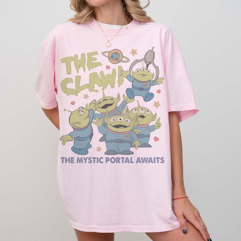 May include: Light pink t-shirt featuring a cartoon graphic of the aliens from the movie Toy Story. The graphic includes the text "THE CLAW!" and "THE MYSTIC PORTAL AWAITS". The aliens are in blue uniforms.