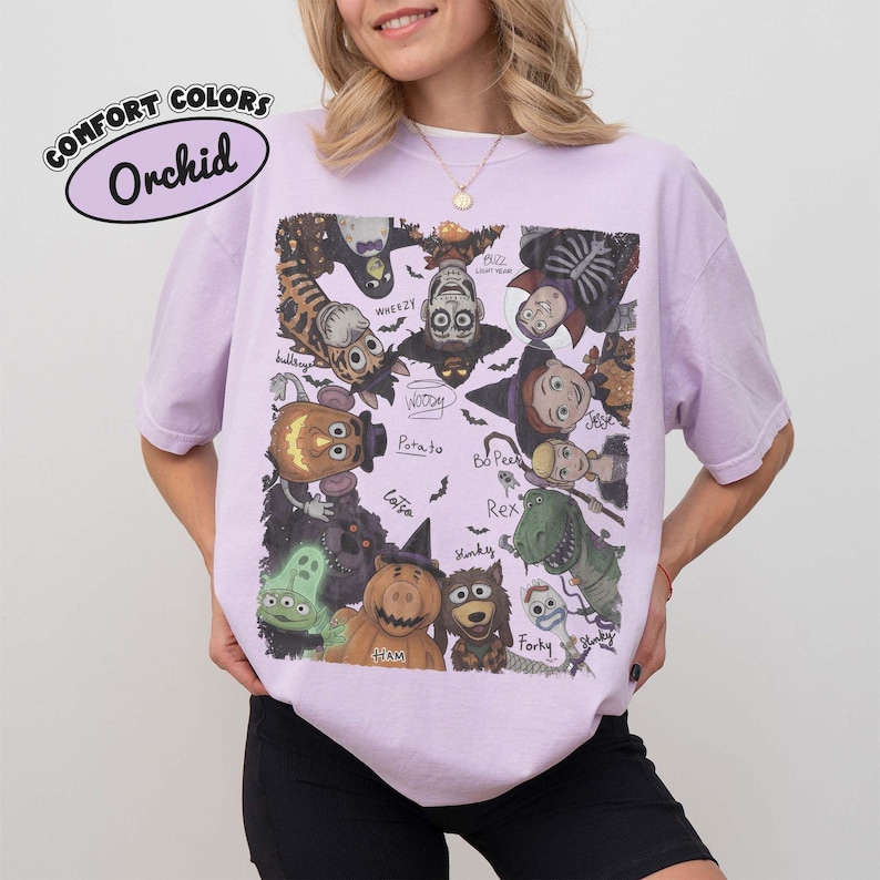 May include: An orchid-colored short-sleeve t-shirt featuring a graphic print of various animated characters dressed in Halloween costumes. The text "COMFORT COLORS" and "Orchid" appears in the upper left corner, with character names labeled.