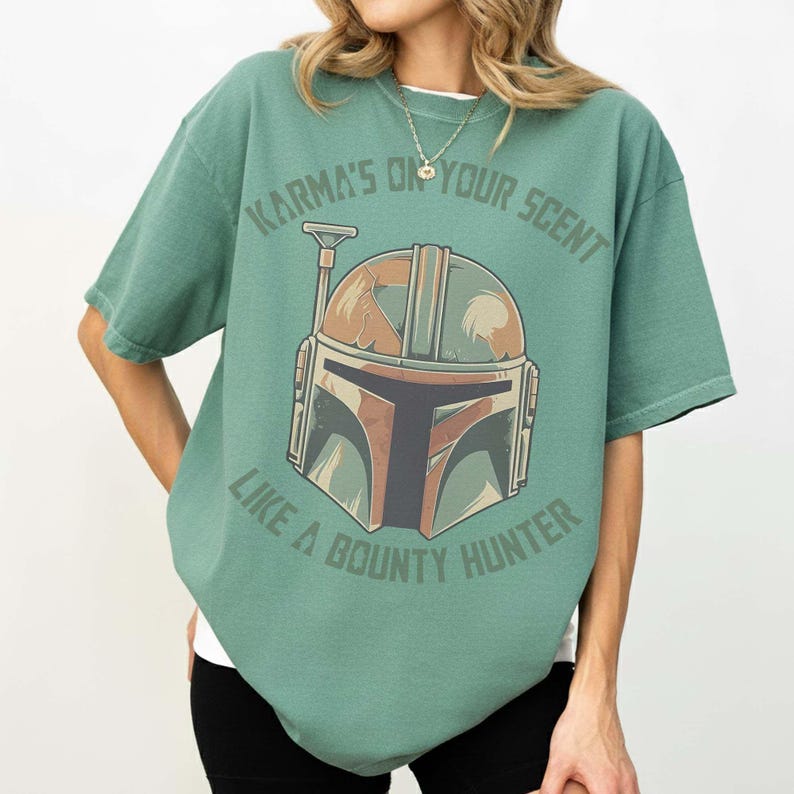 May include: A green t-shirt with a Mandalorian helmet graphic and the text "Karma's on your scent like a bounty hunter." The helmet design incorporates brown, green, and orange hues. The shirt has short sleeves and a loose fit.