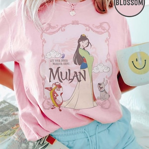 May include: Pink t-shirt featuring a graphic of Mulan with Mushu and a dog. The design includes the text "Let Your Inner Warrior Shine" and "Mulan" in a decorative frame. A mug with a smiley face is held in the hand.