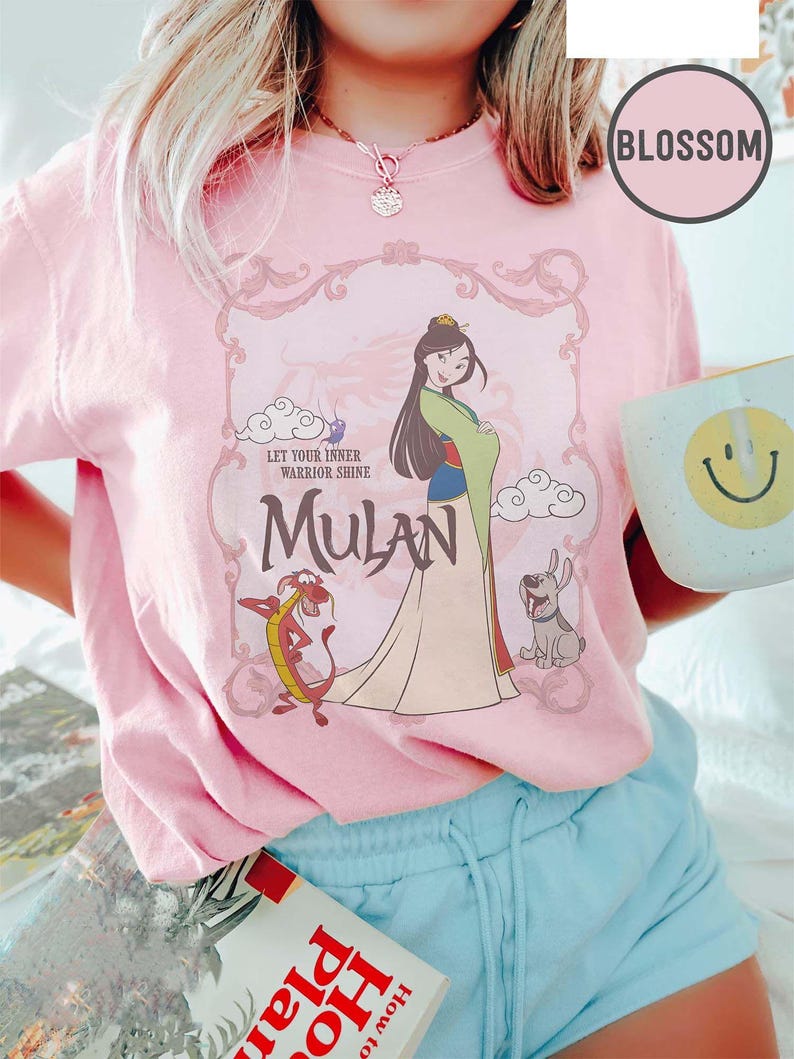 May include: Pink t-shirt featuring a graphic of Mulan with Mushu and a dog. The design includes the text "Let Your Inner Warrior Shine" and "Mulan" in a decorative frame. A mug with a smiley face is held in the hand.