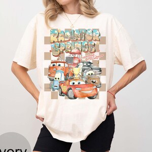 Peut inclure: T-shirt ivoire avec un motif color&eacute; de personnages du film Cars, dont Flash McQueen, et le texte "Radiator Springs". Le motif est plac&eacute; sur un fond &agrave; carreaux.