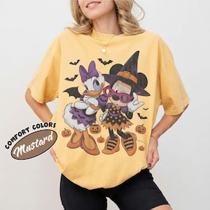 Może przedstawiać: Musztardowy t-shirt z nadrukiem graficznym przedstawiającym Daisy Duck i Minnie Mouse w kostiumach na Halloween. Wz&oacute;r zawiera nietoperze, dynie oraz napis COMFORT COLORS MUSTARD. Koszulka ma kr&oacute;tkie rękawy i okrągły dekolt.