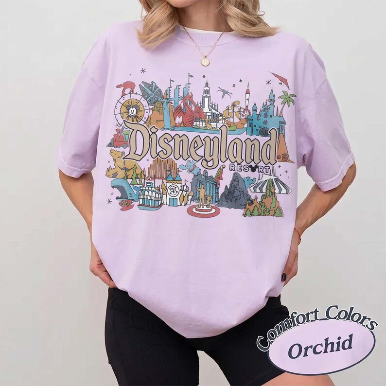 May include: A light purple t-shirt with a Disneyland Resort graphic. The graphic features various park landmarks in a colorful, vintage style. The shirt is labeled "Comfort Colors" and "Orchid".
